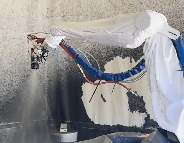 A cobot in a white protective cover spraying a grey coating onto a test wall with an automatic spray gun